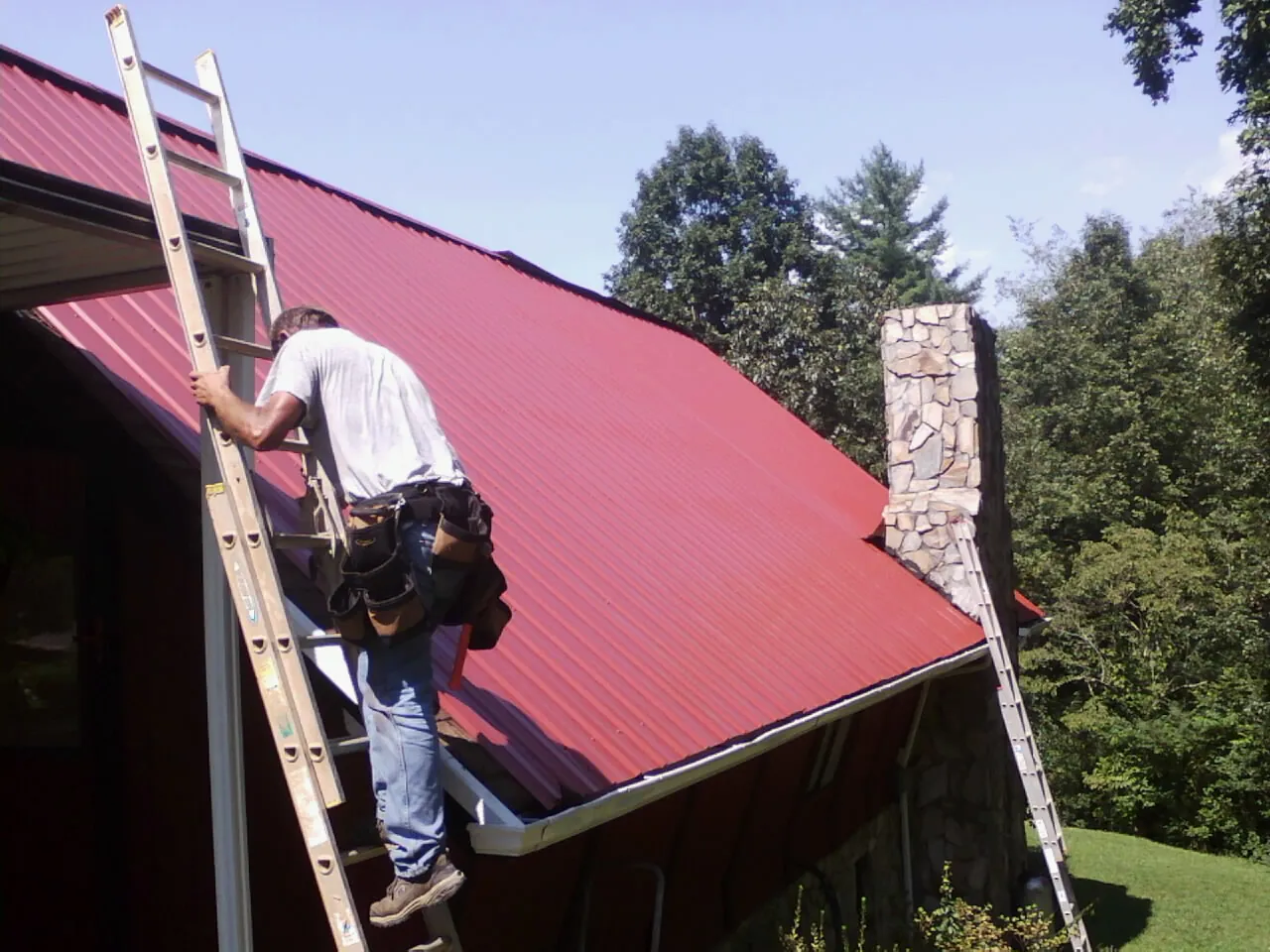 Roofer on a ladder beside a red metal roof and stone chimney — Soffit & Fascia Repair by Roofing Brothers in Anna